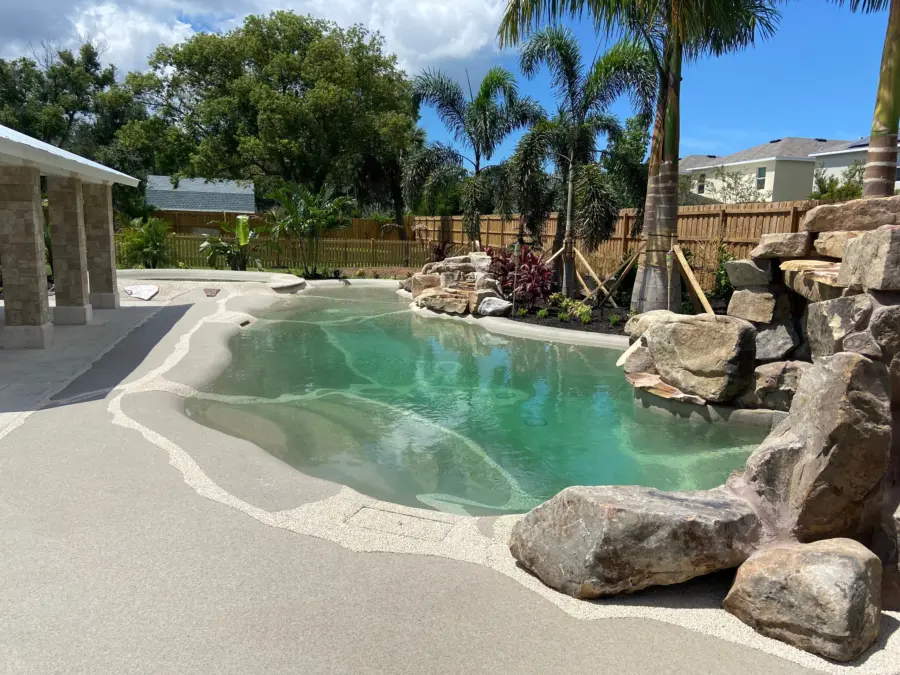 Beach-entry pool with sand-colored deck, boulder outcrops, palms and stone-column pavilion