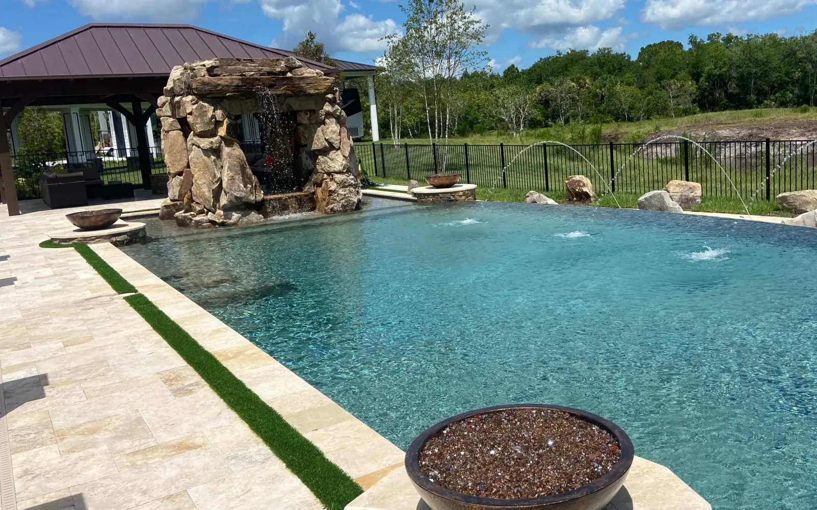 Pool with boulder grotto waterfall, copper-tone fire bowls, deck jets, travertine deck and pavilion