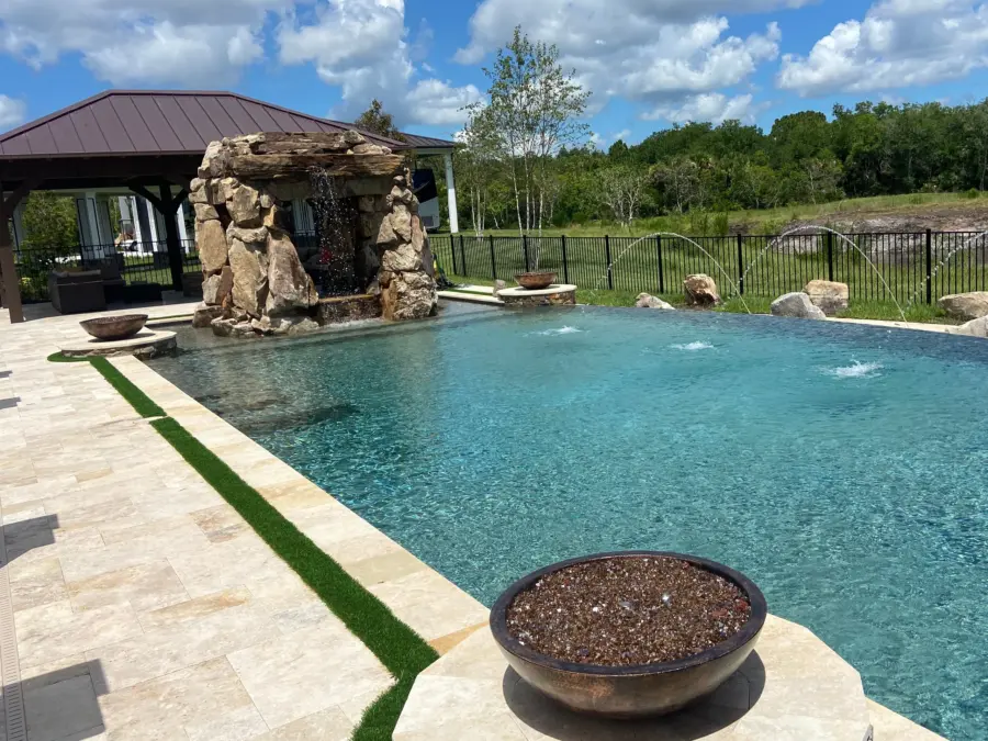 Pool with boulder grotto waterfall, copper-tone fire bowls, deck jets, travertine deck and pavilion