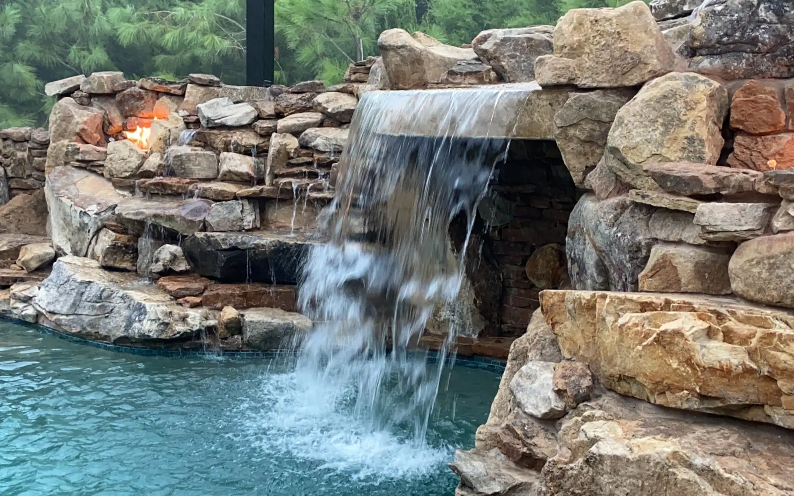 Close view of a boulder grotto waterfall pouring into a pool between two lit fire niches