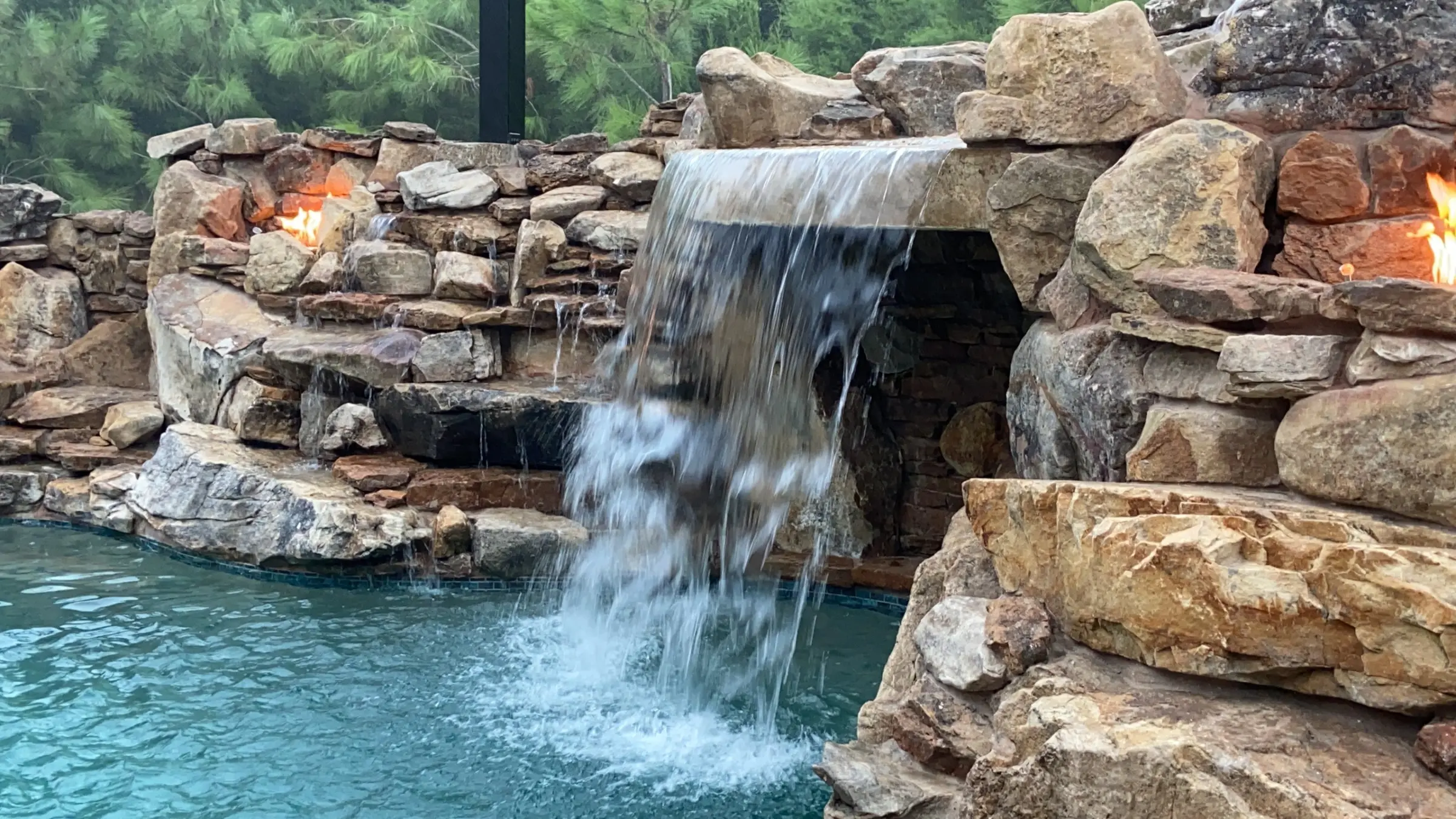 Close view of a boulder grotto waterfall pouring into a pool between two lit fire niches