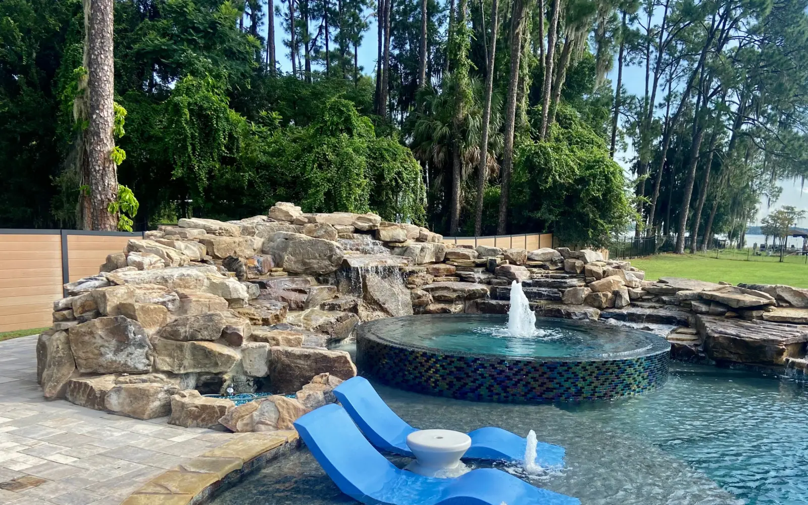 Boulder waterfall beside a raised round spa with glass tile, sun shelf loungers and bubblers