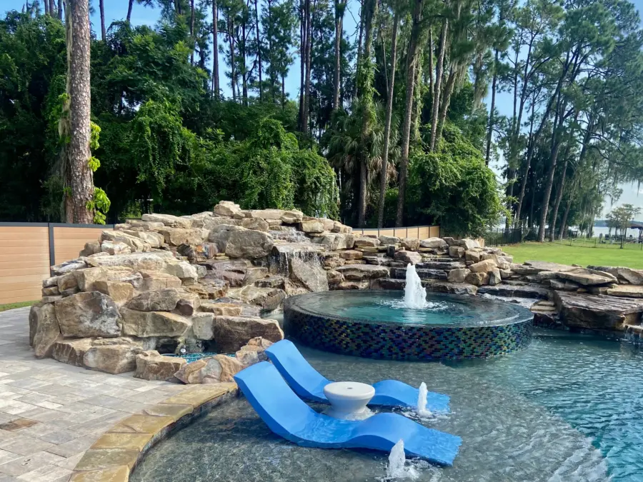 Boulder waterfall beside a raised round spa with glass tile, sun shelf loungers and bubblers