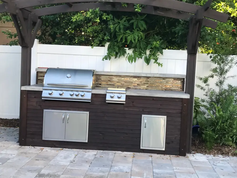 Compact outdoor kitchen with dark stained wood cabinet, gray countertop, glass tile backsplash and Blaze grill