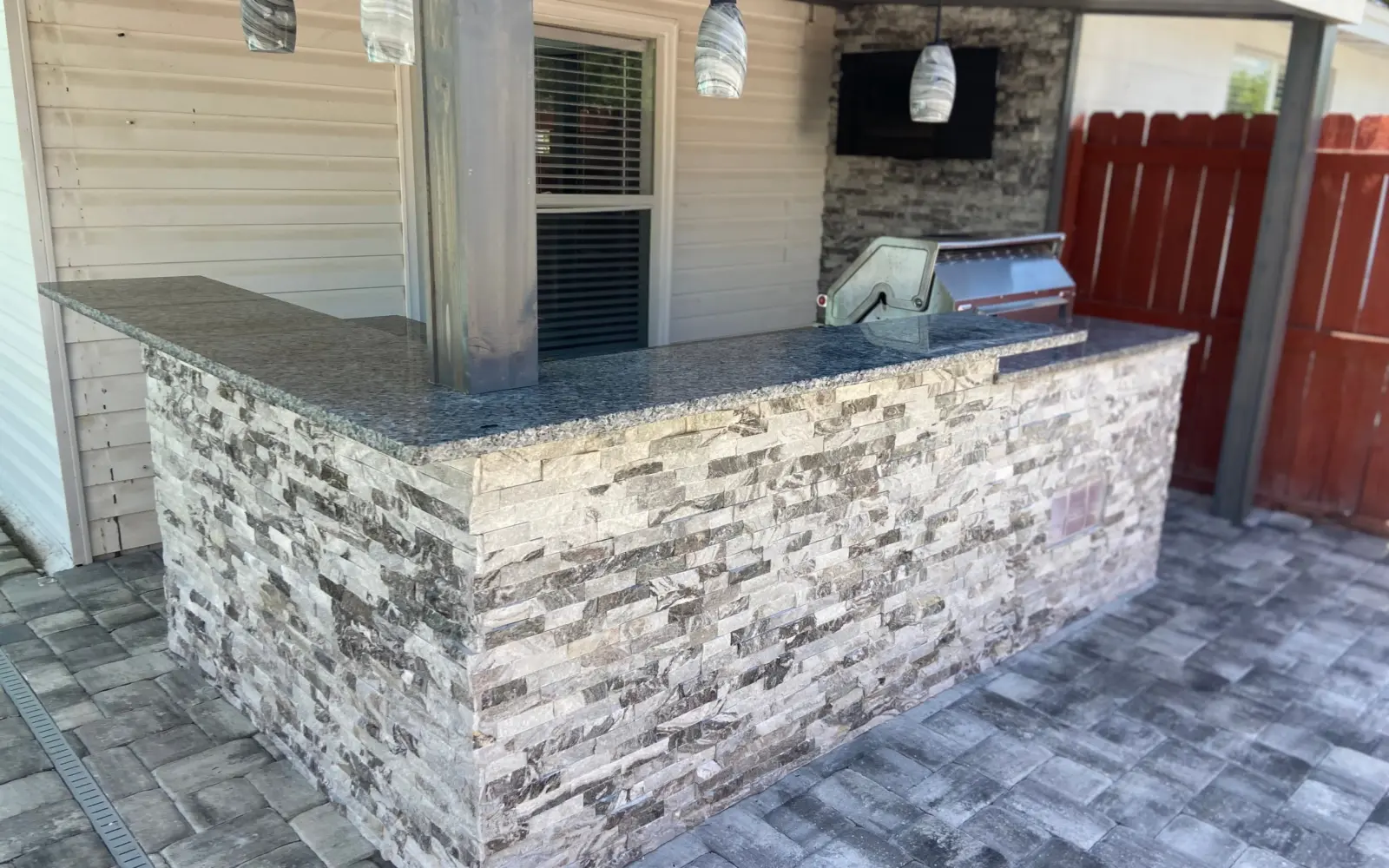 L-shaped outdoor bar in gray stacked stone with dark stone countertop, pendant lights and TV