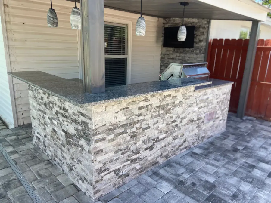 L-shaped outdoor bar in gray stacked stone with dark stone countertop, pendant lights and TV