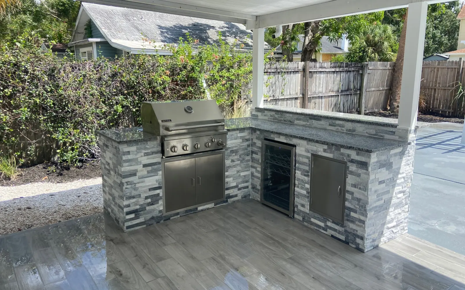 L-shaped outdoor kitchen in gray stacked stone with a built-in grill, wine fridge and stainless door