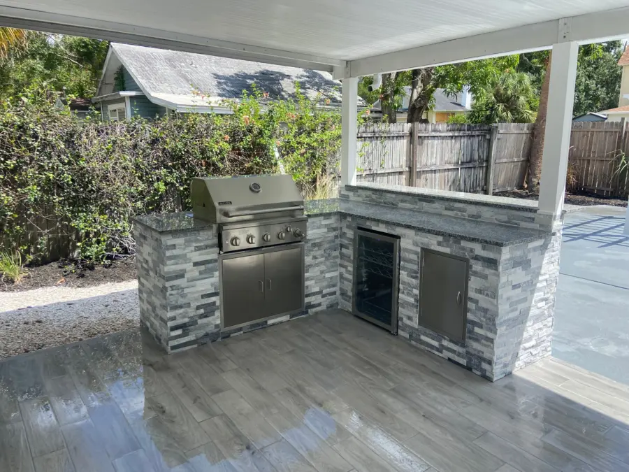 L-shaped outdoor kitchen in gray stacked stone with a built-in grill, wine fridge and stainless door