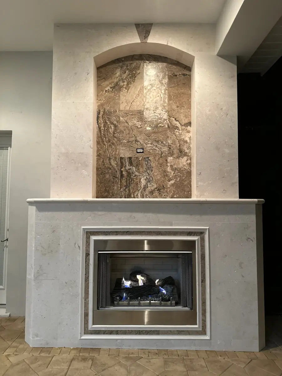 Lit lanai gas fireplace with cream stone surround and arched brown veined stone niche at dusk