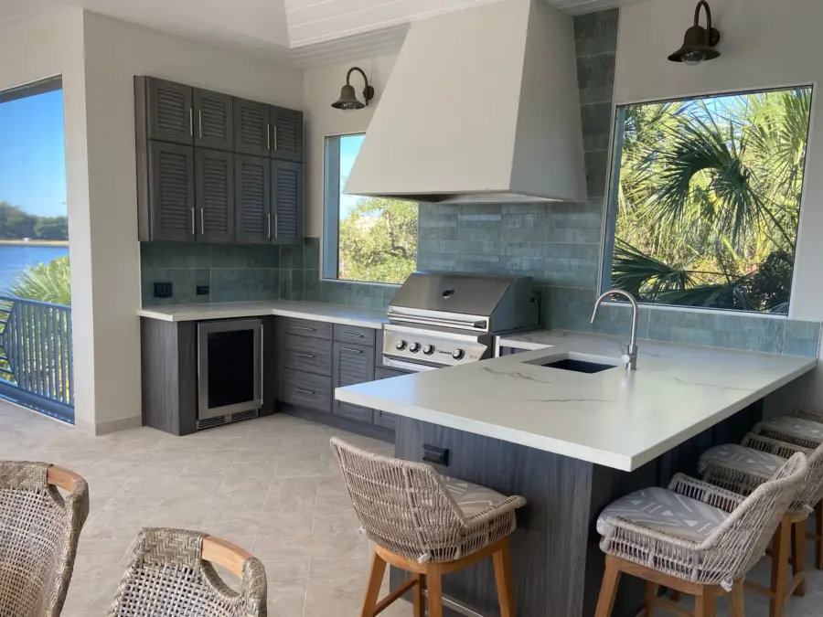 Outdoor kitchen on a lanai with gray louvered cabinets, white veined countertop and plaster vent hood