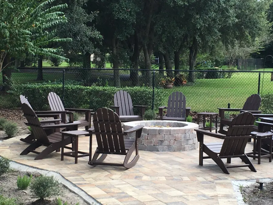 Round paver patio with block fire pit ringed by dark Adirondack chairs and side tables