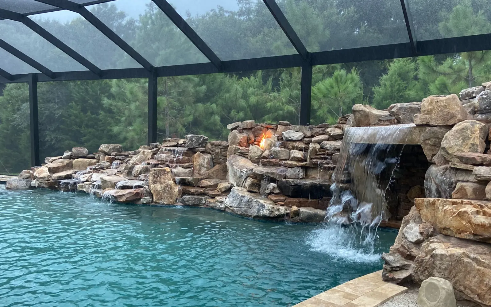 Screened pool with boulder grotto waterfall, lit fire niches in the rockwork and travertine coping