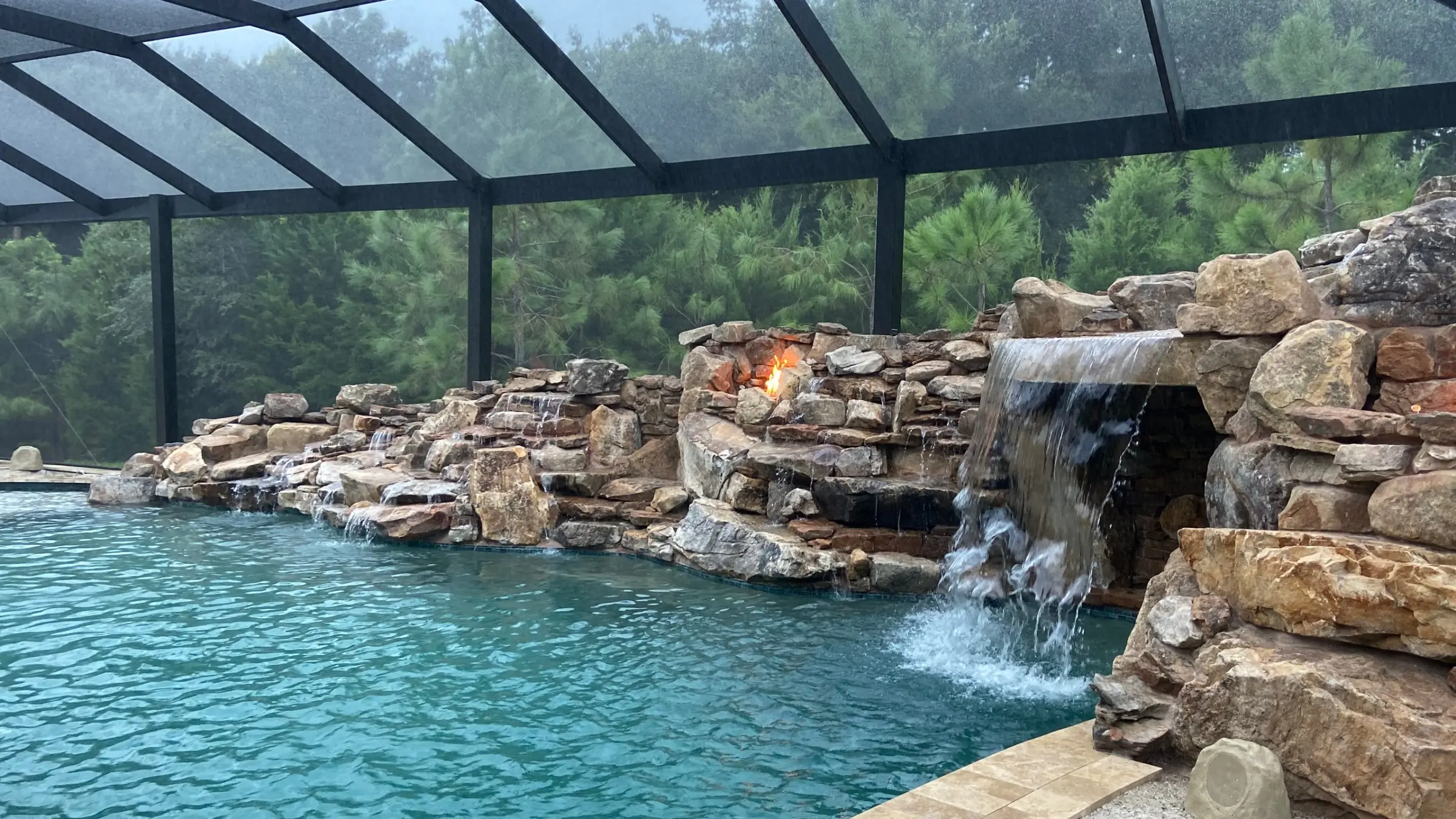 Screened pool with boulder grotto waterfall, lit fire niches in the rockwork and travertine coping
