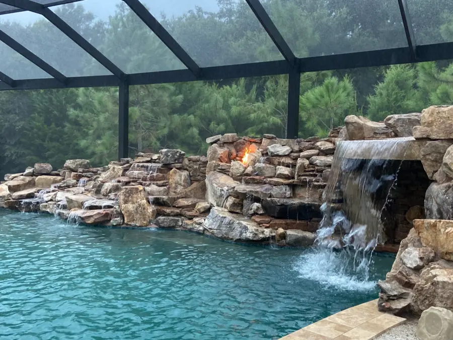 Screened pool with boulder grotto waterfall, lit fire niches in the rockwork and travertine coping