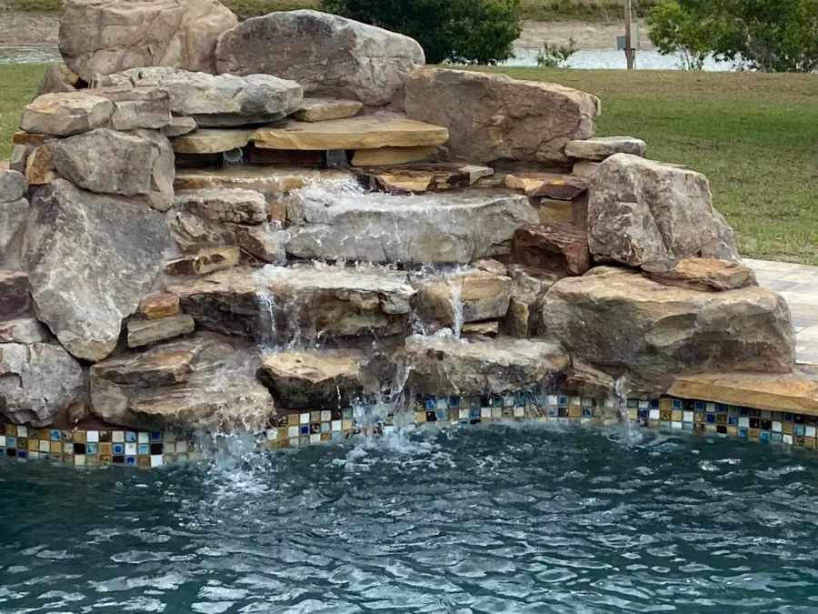 Stacked boulder waterfall spilling over flat ledges into a pool with glass mosaic tile