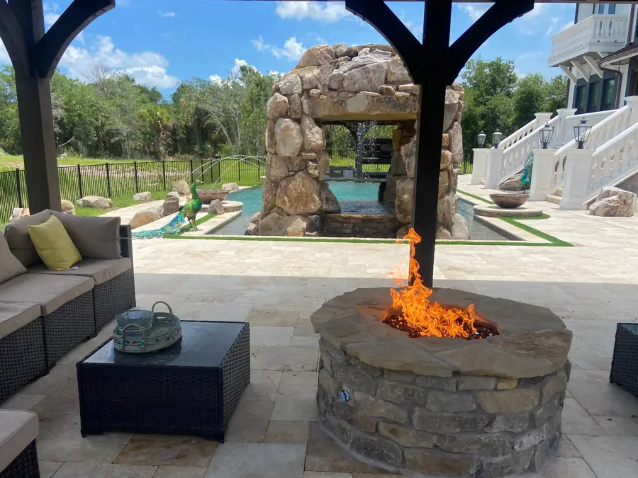 Round stone gas fire pit with flagstone cap burning under a timber pavilion, boulder grotto beyond