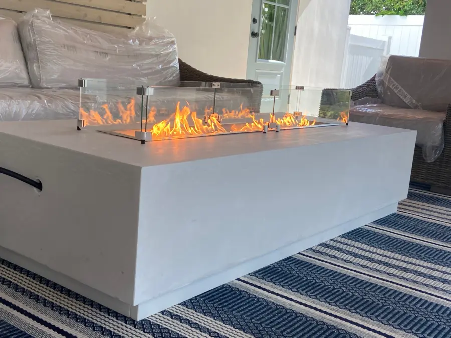 White linear fire table with glass wind guard and lit gas flames on a covered patio