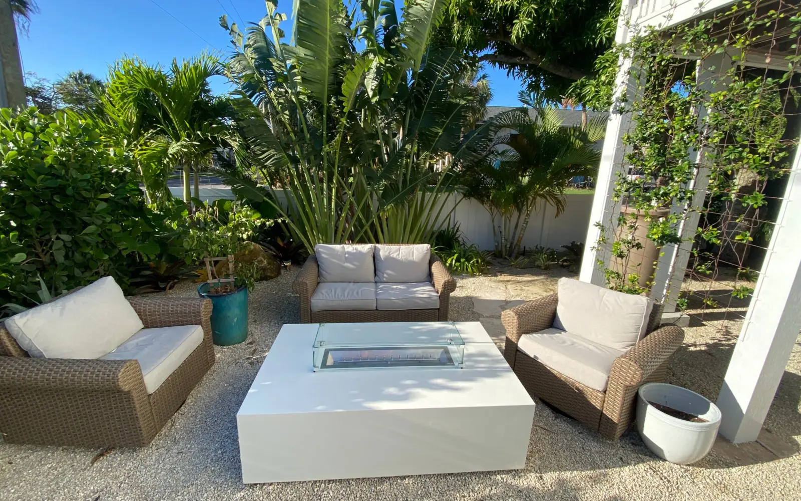 White rectangular fire table with glass wind guard among wicker seating in a gravel garden