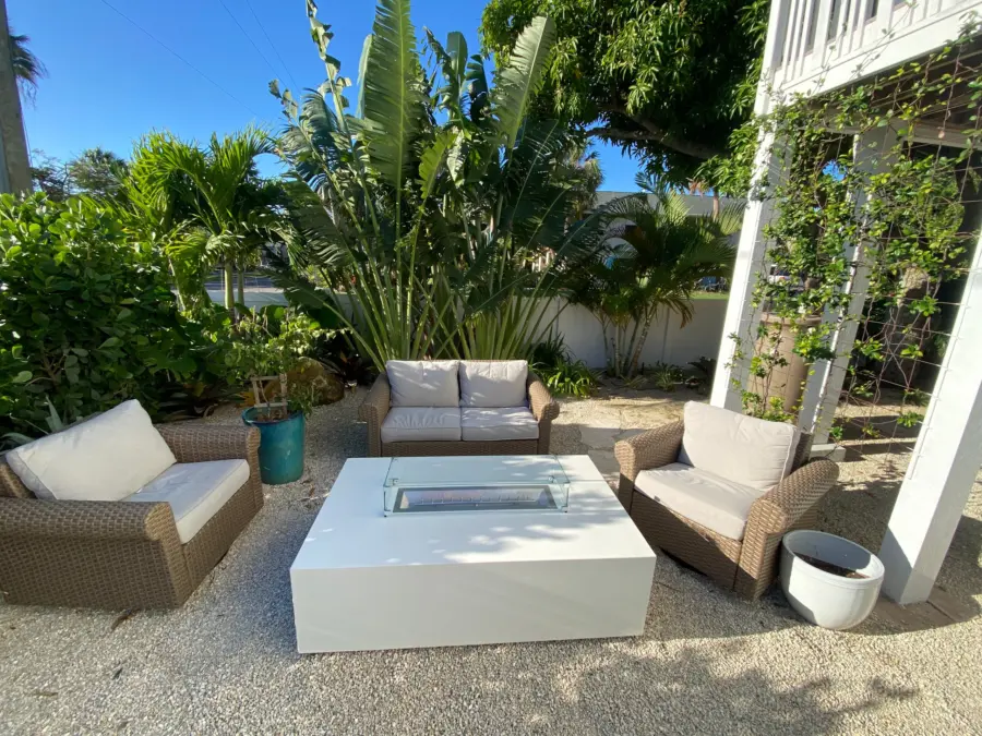 White rectangular fire table with glass wind guard among wicker seating in a gravel garden